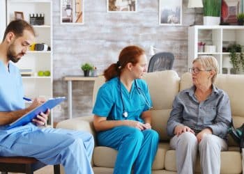 Equipe multiprofissional da área da saúde atende uma paciente idosa em casa, representando as áreas de atuação da Enfermagem e Psicologia.