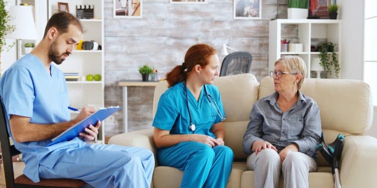 Equipe multiprofissional da área da saúde atende uma paciente idosa em casa, representando as áreas de atuação da Enfermagem e Psicologia.