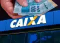 Mão segurando cédulas de R$ 100 em frente à fachada azul da Caixa Econômica Federal, ilustrando a liberação do saque calamidade do FGTS para novos municípios em abril de 2026.
