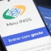 Tela de login do aplicativo Meu INSS aberta em um celular sobre teclado de notebook, ao lado de cartão da Previdência Social.