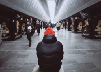 Pessoa de touca laranja e casaco escuro sentada de costas em corredor movimentado de estação, simbolizando a sensação de solidão em meio à multidão