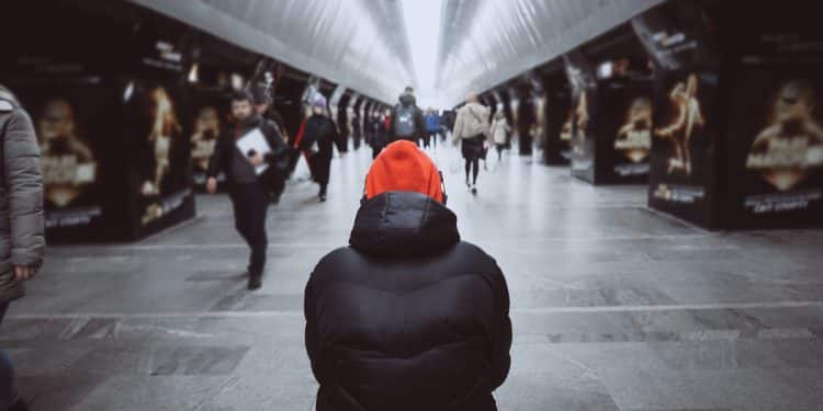 Pessoa de touca laranja e casaco escuro sentada de costas em corredor movimentado de estação, simbolizando a sensação de solidão em meio à multidão