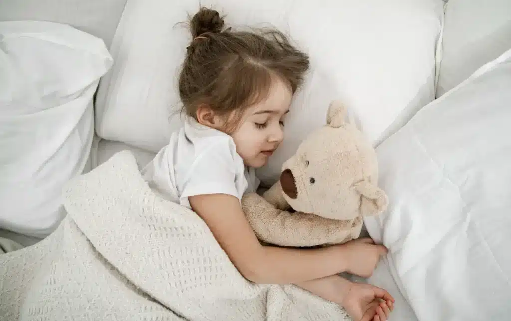 Criança pequena dormindo com ursinho na cama representando a importância do sono na fase pré-escolar