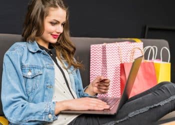 Mulher realizando compra online com cartão de crédito em notebook, cercada por sacolas de compras.