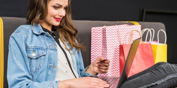 Mulher realizando compra online com cartão de crédito em notebook, cercada por sacolas de compras.