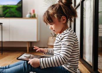 Menina de blusa listrada sentada no chão da sala utilizando tablet sozinha enquanto televisão passa imagens ao fundo