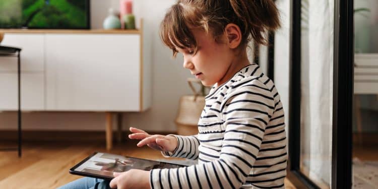 Menina de blusa listrada sentada no chão da sala utilizando tablet sozinha enquanto televisão passa imagens ao fundo