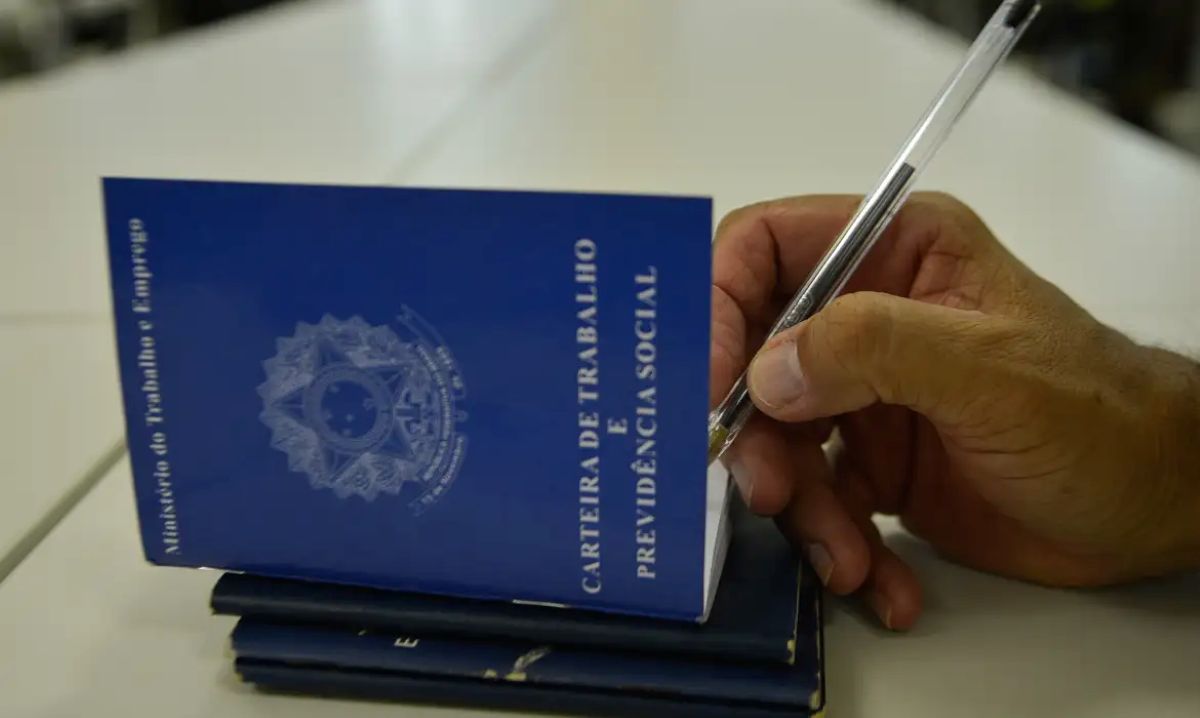 Close de uma mão masculina segurando uma caneta sobre uma Carteira de Trabalho e Previdência Social (CTPS) azul aberta, com outras carteiras empilhadas abaixo sobre uma mesa branca.