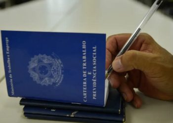 Close de uma mão masculina segurando uma caneta sobre uma Carteira de Trabalho e Previdência Social (CTPS) azul aberta. A mão está prestes a assinar ou preencher o documento sobre uma mesa branca.