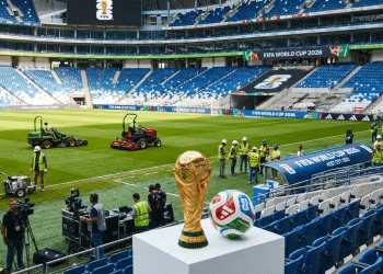Troféu dourado da Copa do Mundo FIFA 2026 e bola oficial em pedestal branco com trabalhadores no gramado do estádio ao fundo