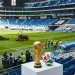Troféu dourado da Copa do Mundo FIFA 2026 e bola oficial em pedestal branco com trabalhadores no gramado do estádio ao fundo