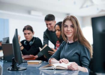 Vista em primeiro plano de uma estudante jovem com sardas e cabelo castanho longo, usando suéter cinza e camisa de gola rosa. Ela sorri para a câmera enquanto lê um livro aberto em uma mesa de laboratório de informática. Ao fundo, outros três estudantes (dois homens e uma mulher) estão desfocados, interagindo com livros e computadores.