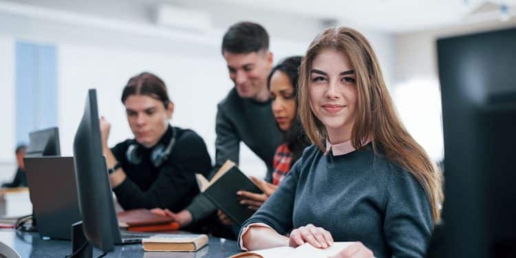 Vista em primeiro plano de uma estudante jovem com sardas e cabelo castanho longo, usando suéter cinza e camisa de gola rosa. Ela sorri para a câmera enquanto lê um livro aberto em uma mesa de laboratório de informática. Ao fundo, outros três estudantes (dois homens e uma mulher) estão desfocados, interagindo com livros e computadores.