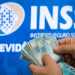 Mãos contando notas de R$ 100 com logo do INSS e o texto Previdência Social ao fundo, em painel azul, simbolizando o pagamento da segunda parcela do 13º salário e os golpes a ela relacionados.