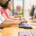 Três pessoas sentadas em fila digitando em teclados pretos diante de monitores em mesa de madeira, simbolizando os cursos livres.