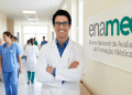 Médico sorridente diante de banner do ENAMED em corredor hospitalar.