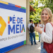 Jovem loira com mochila vermelha faz sinal positivo ao lado de banner do programa Pé-de-Meia em pátio escolar.