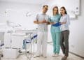 Dentista, assistente de uniforme azul-turquesa e mulher de camisa azul-clara analisam prancheta em consultório odontológico, simbolizando o novo piso salarial de médicos e cirurgiões-dentistas.
