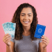Jovem sorridente segura notas de 100 reais e Carteira de Trabalho e Previdência Social azul em fundo rosa.