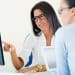 Duas mulheres em escritório, uma de blusa branca aponta para monitor enquanto outra de camisa azul observa atentamente, simbolizando a diferença entre RH e Departamento pessoal.