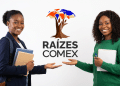Duas jovens negras sorrindo e apontando para logo do Raízes Comex, uma com blazer e livros, outra com blusa verde e caderno.