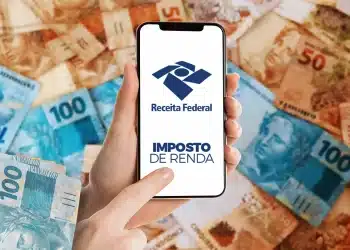 Mão segurando smartphone com tela da Receita Federal sobre cédulas brasileiras de 50 e 100 reais.