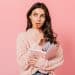 Mulher jovem de suéter rosa tricotado segura caderno e lápis vermelho enquanto olha pensativa para o lado, simbolizando a dúvida clássica entre " a gente vai" e " a gente vamos".