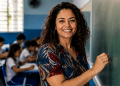 Professora sorri ao lado do quadro em uma sala de aula com alunos ao fundo