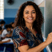 Professora sorri ao lado do quadro em uma sala de aula com alunos ao fundo