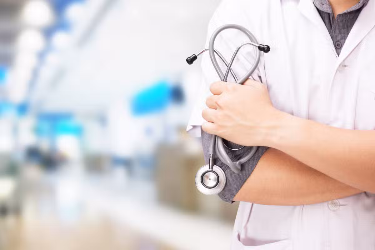 Médico de jaleco branco, de braços cruzados, segurando um estetoscópio em corredor de hospital, ilustrando a divulgação do resultado dos recursos do atendimento especializado do Enamed 2026