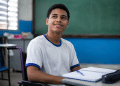 Jovem cadeirante com camiseta branca de gola azul sorri em sala de aula com lousa verde ao fundo.