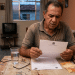 Homem idoso lê documento oficial com expressão preocupada à mesa da sala de casa