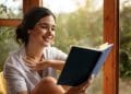 Mulher sorridente lendo livro azul próxima a uma janela com vegetação ao fundo