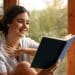 Mulher sorridente lendo livro azul próxima a uma janela com vegetação ao fundo