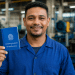Trabalhador de uniforme azul sorri segurando a Carteira de Trabalho e Previdência Social dentro de uma fábrica