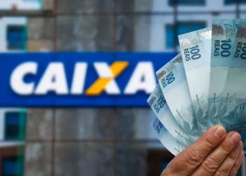 Mão segurando notas de R$ 100 em frente a uma agência da CAIXA, destacando o valor de R$ 6.220 disponível para brasileiros.
