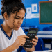 Jovem estudante com uniforme branco e azul concentrada usando calculadora científica em sala de aula com lousa verde.