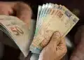 Duas mãos segurando maços de cédulas brasileiras de 50 e 20 reais em leque, fundo escuro desfocado, simbolizando o pagamento do Abono Salarial 2026.