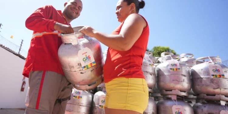 Homem de uniforme vermelho entregando botijão de gás identificado com etiqueta do programa para uma mulher em ponto de distribuição