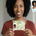 Mulher negra sorridente com cabelo crespo mostra a Carteira de Identidade Nacional emitida na Bahia, simbolizando a ação de emissão da Nova Carteira de Identidade em Salvador.