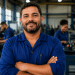 Trabalhador de uniforme azul sorri de braços cruzados em uma fábrica, com colegas ao fundo