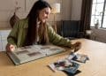 Jovem mulher sorridente sentada à mesa folheando álbum de fotografias antigas com expressão tranquila