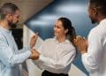 Mulher de camisa branca cumprimenta colega enquanto outro aplaude em ambiente corporativo com parede azul, simbolizando os comportamentos de quem teve muitos amigos na infância.