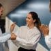 Mulher de camisa branca cumprimenta colega enquanto outro aplaude em ambiente corporativo com parede azul, simbolizando os comportamentos de quem teve muitos amigos na infância.