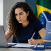 Mulher de cabelos cacheados escrevendo documento com caneta, laptop aberto, placa Câmara Municipal e bandeira do Brasil ao fundo.