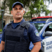 Guarda Municipal de uniforme azul marinho com colete balístico e rádio comunicador, diante de viatura oficial.