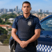 Agente da Guarda Municipal de uniforme azul-marinho com colete balístico ao lado de viatura com brasão da corporação.