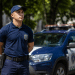 Agente da Guarda Municipal com uniforme azul-marinho e boné ao lado de viatura com inscrição "Guarda Municipal".