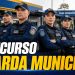 Sete guardas municipais uniformizados com coletes táticos azuis, bonés e distintivos posicionados em frente à viatura e base operacional.