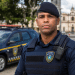 Agente da Guarda Municipal com uniforme azul e colete, rádio comunicador, em frente a viatura e igreja colonial.